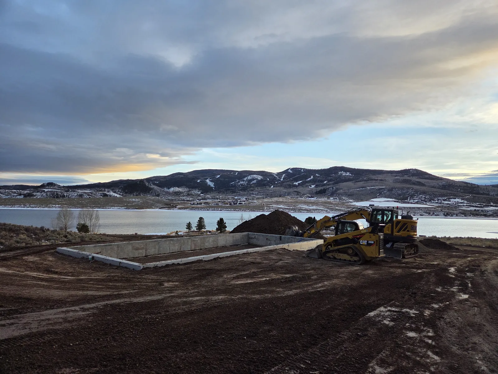 Excavator and skid steer beside a new foundation at dawn, a reservoir and snow-dusted hills behind
