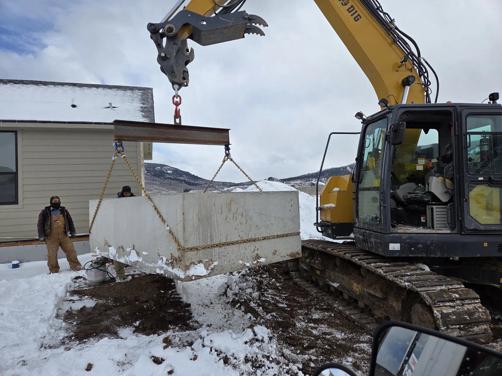 Excavator lifting a concrete septic tank on chains while a crew member watches
